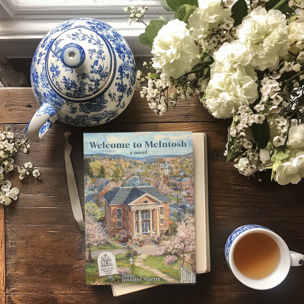 Flatlay photo of Welcome to McIntosh novel with a cup of tea, white flowers, and a blue and white teapot on a rustic table.