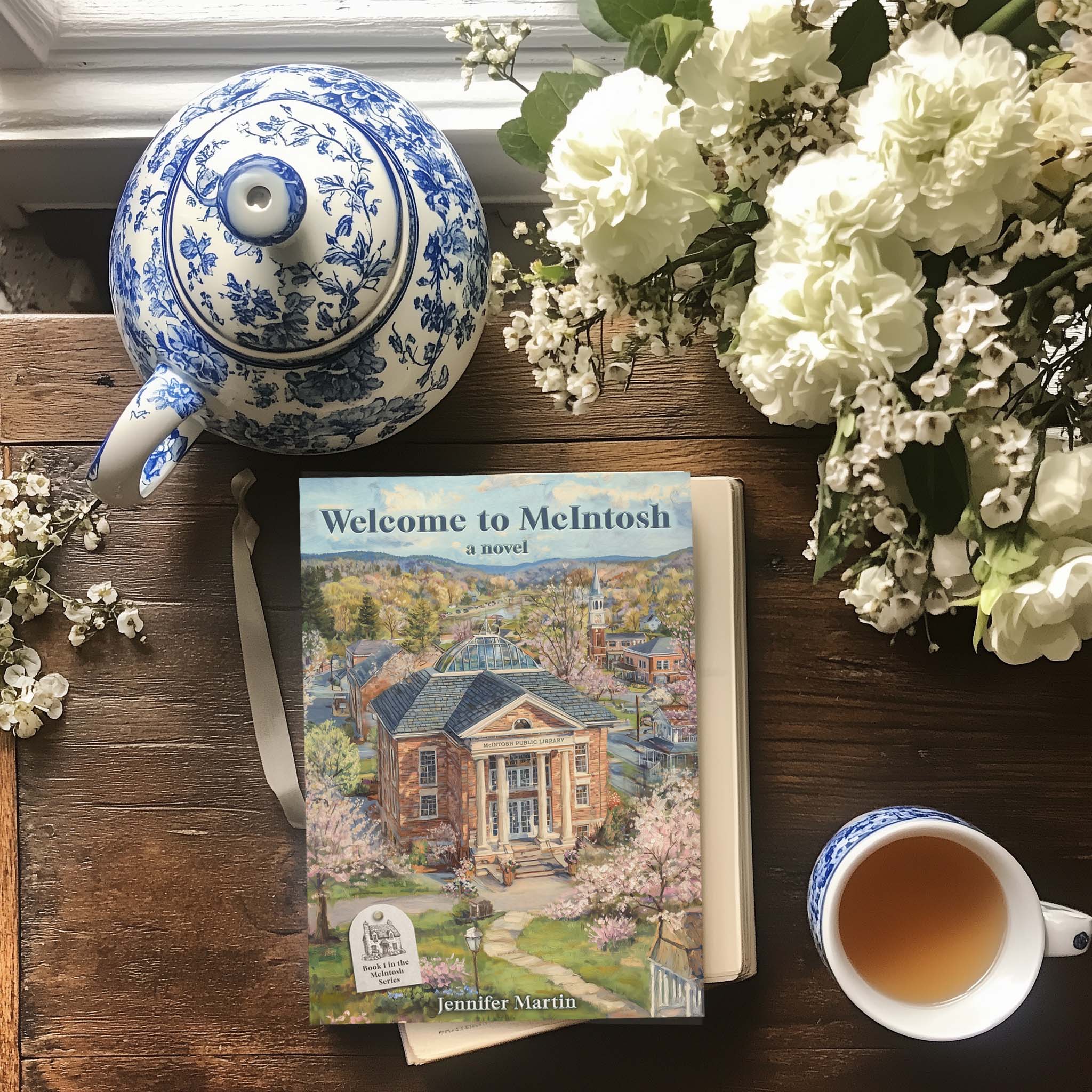 Flatlay photo of Welcome to McIntosh novel with a cup of tea, white flowers, and a blue and white teapot on a rustic table.
